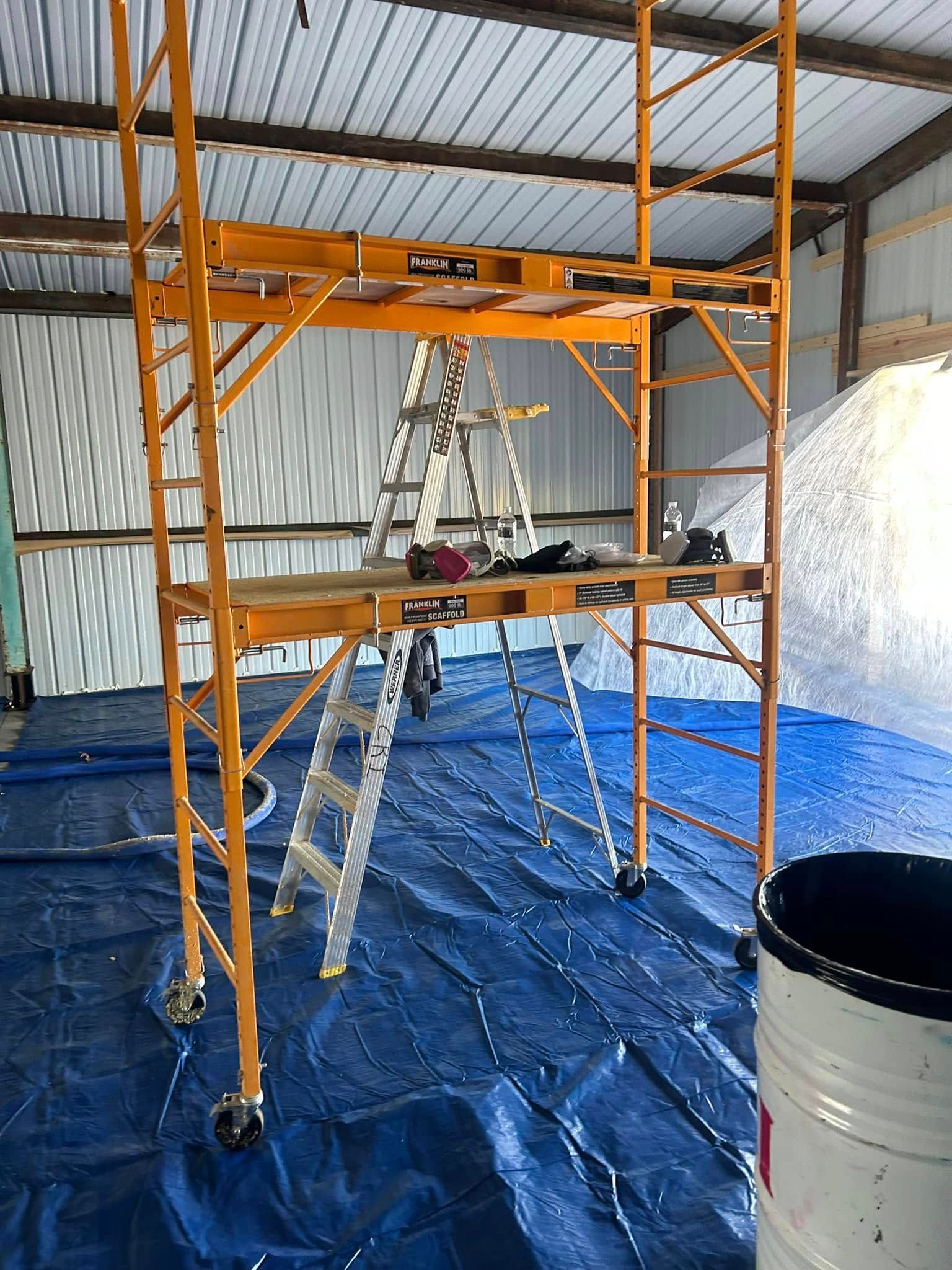 Yellow scaffolding with a ladder inside set up on a blue tarp inside a building. A white bucket sits nearby.