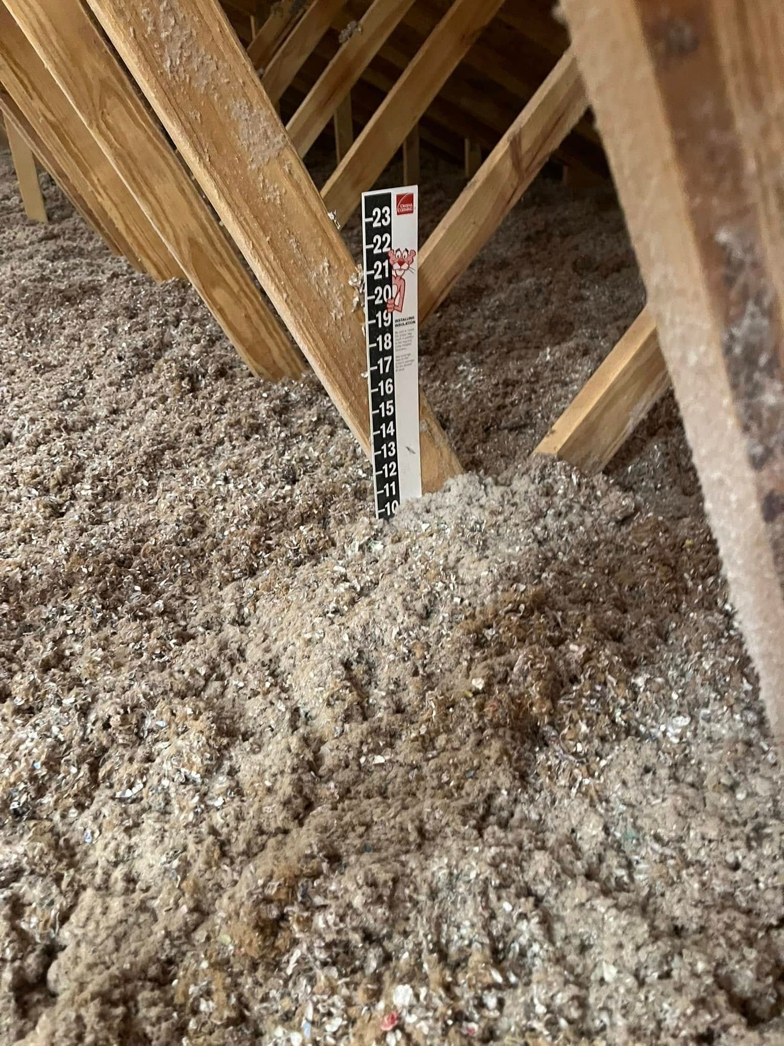 Attic filled with loose-fill insulation, with a ruler measuring its depth between wooden rafters.