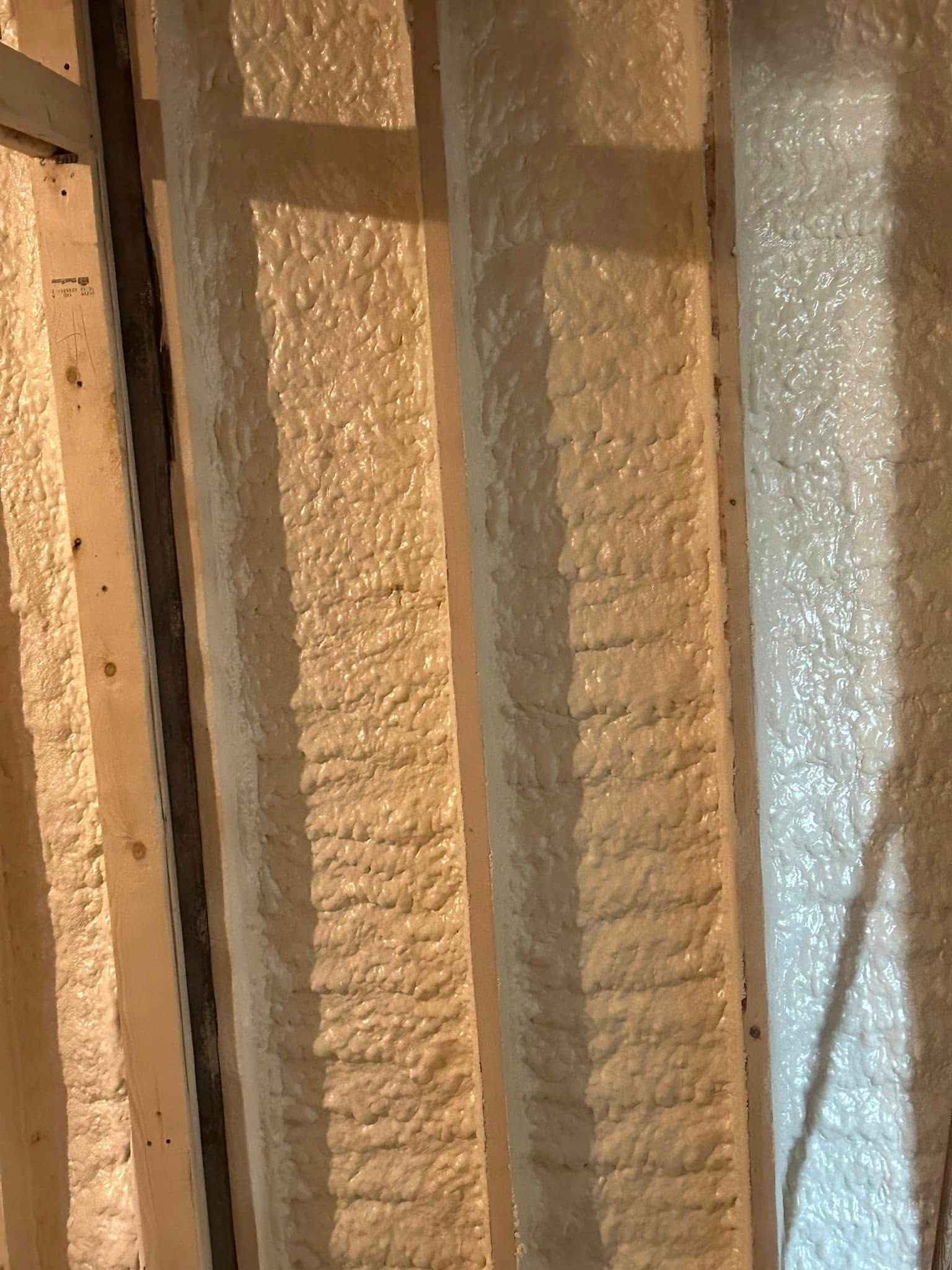 Spray foam insulation fills the space between wooden wall studs, appearing bumpy and off-white.