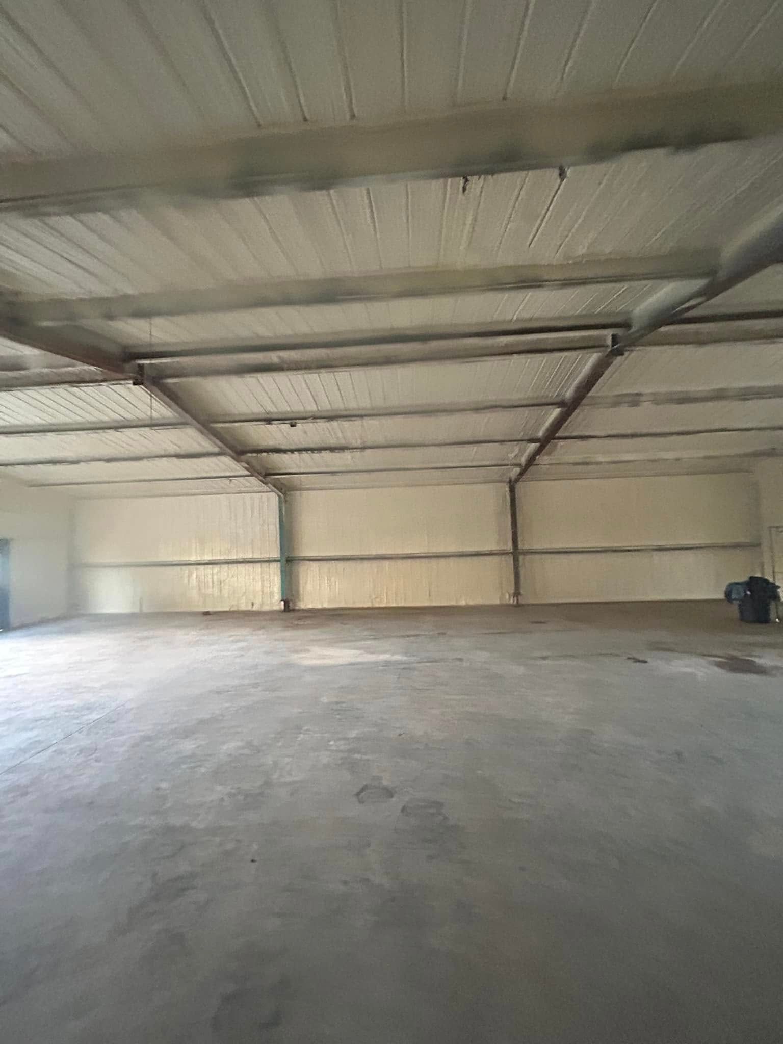 Empty, large warehouse space with concrete floor, corrugated metal roof, and light-colored walls.