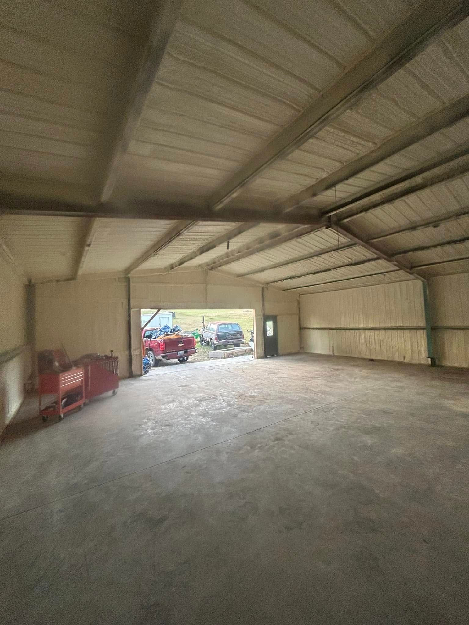 Interior of a large, open garage or shed with a concrete floor and spray foam insulation on the walls and ceiling. The open door reveals vehicles outside.