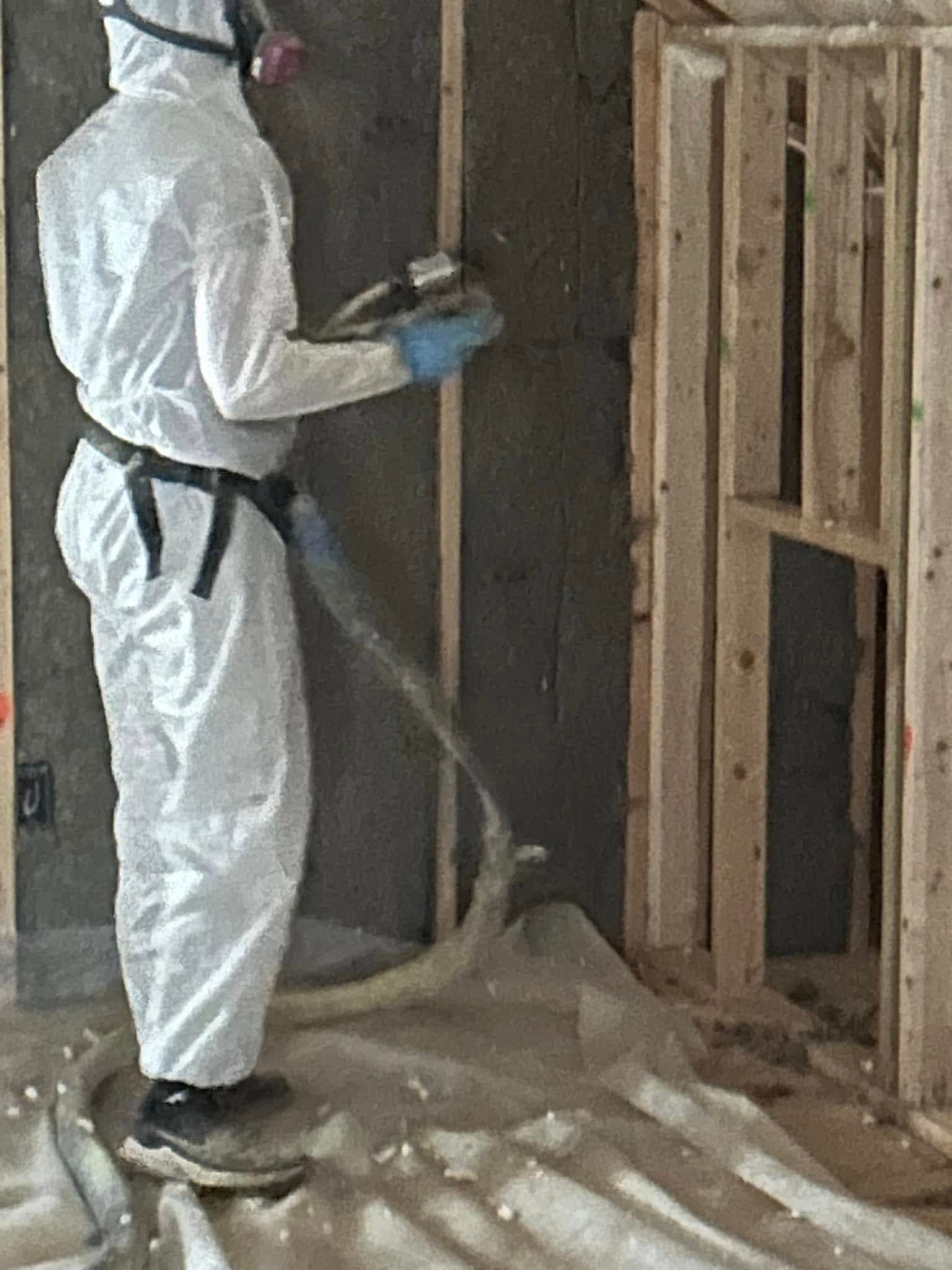 Person in protective suit spraying insulation into a wall. They wear a mask and gloves, standing in a room with wood framing.