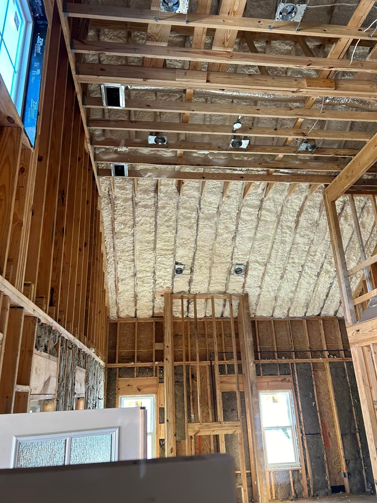 Interior view of a house under construction, showing wooden framing, insulation sprayed on the ceiling, and visible vents.