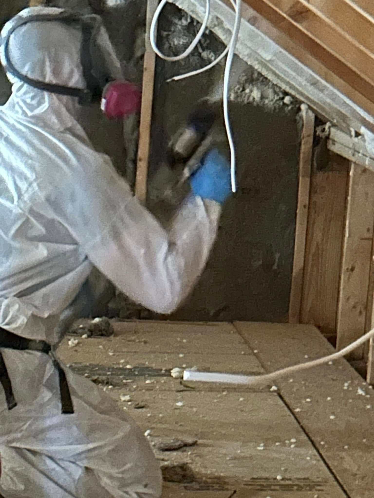 Person in white protective suit and respirator sprays insulation into attic wall.