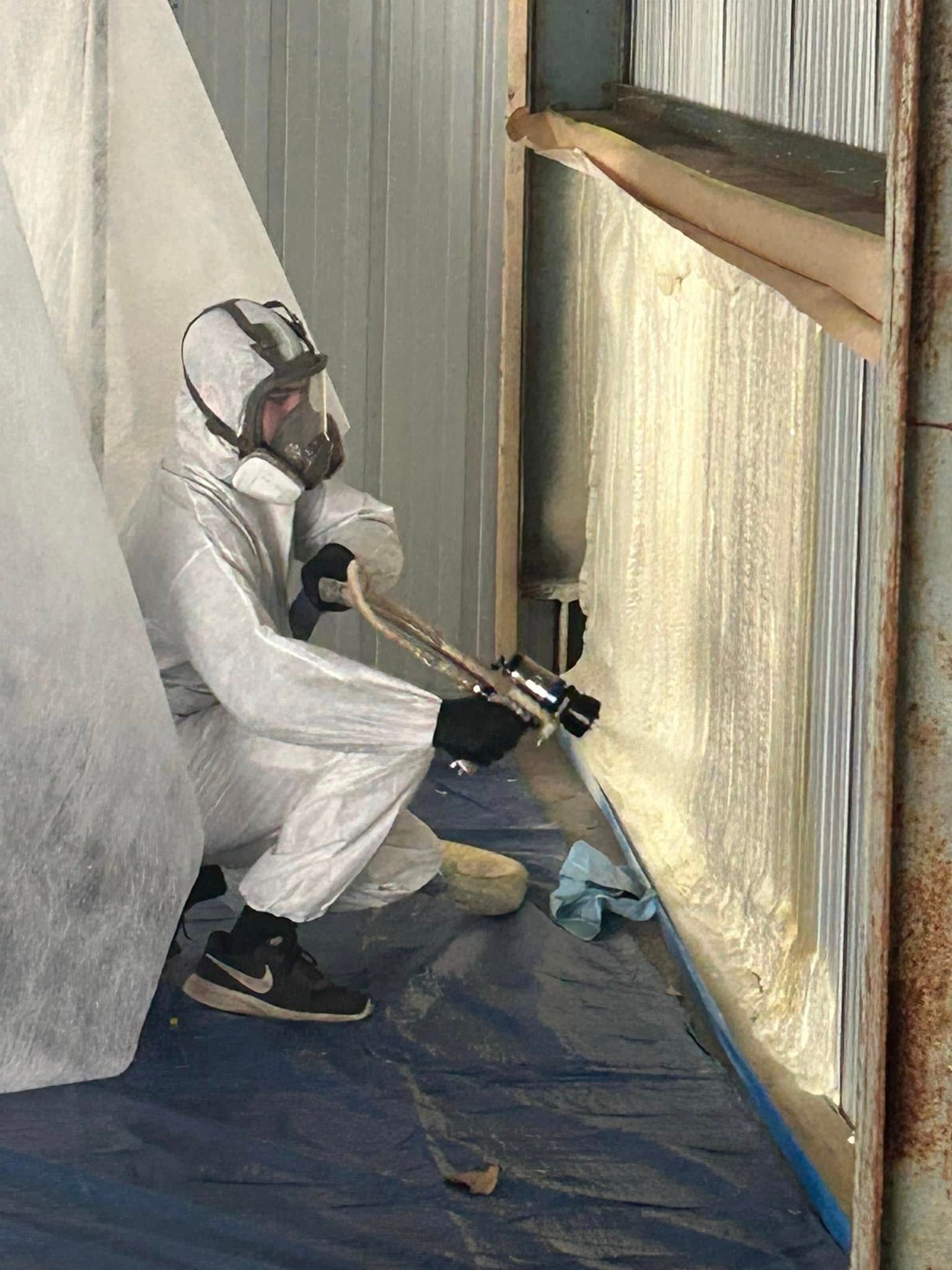Person in protective suit spraying a wall with foam. They are in an enclosed area, with a tarp on the floor.