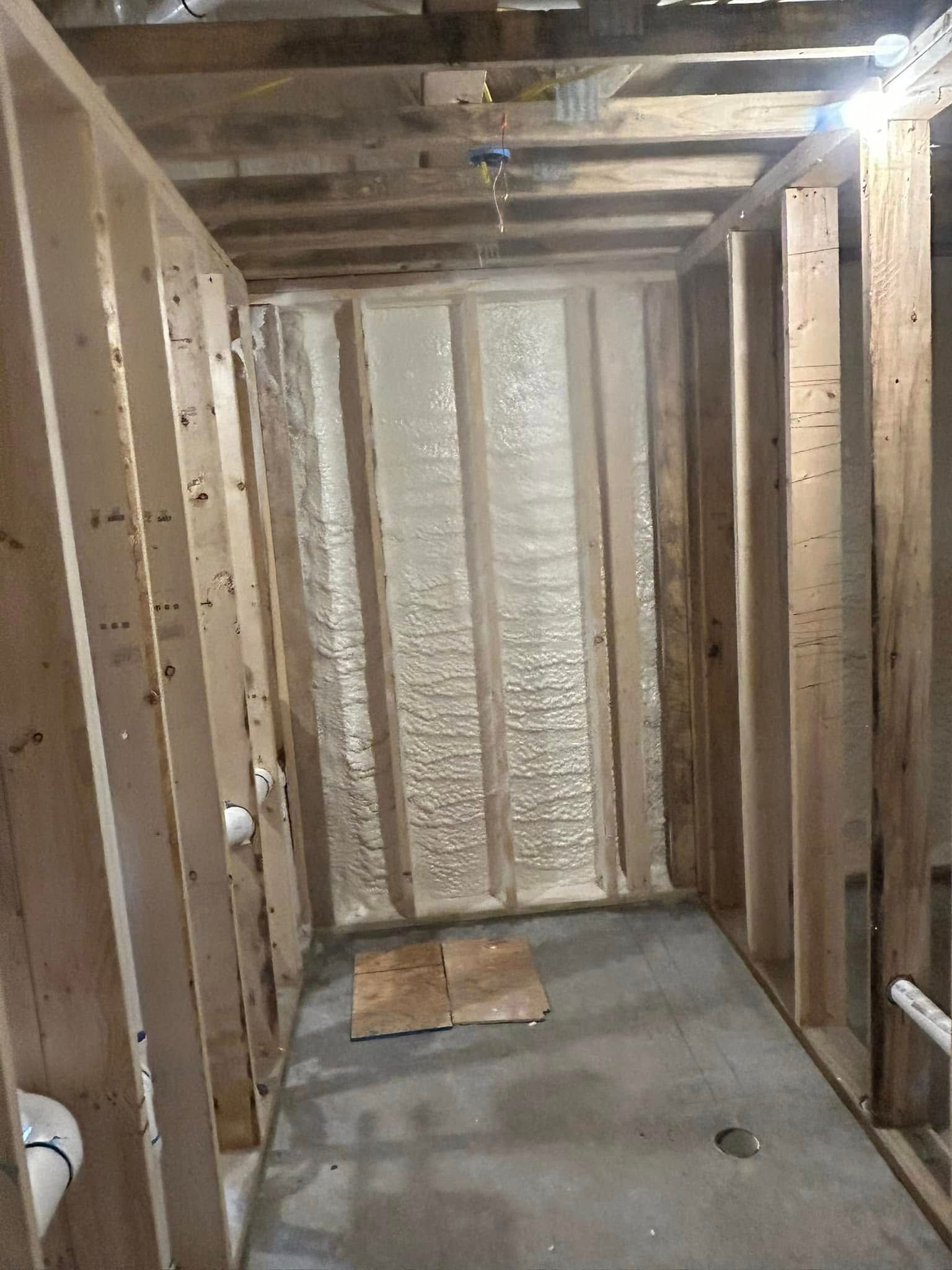 Interior view of a bathroom under construction. Wooden frame walls surround a concrete floor, with spray foam insulation visible on the back wall.