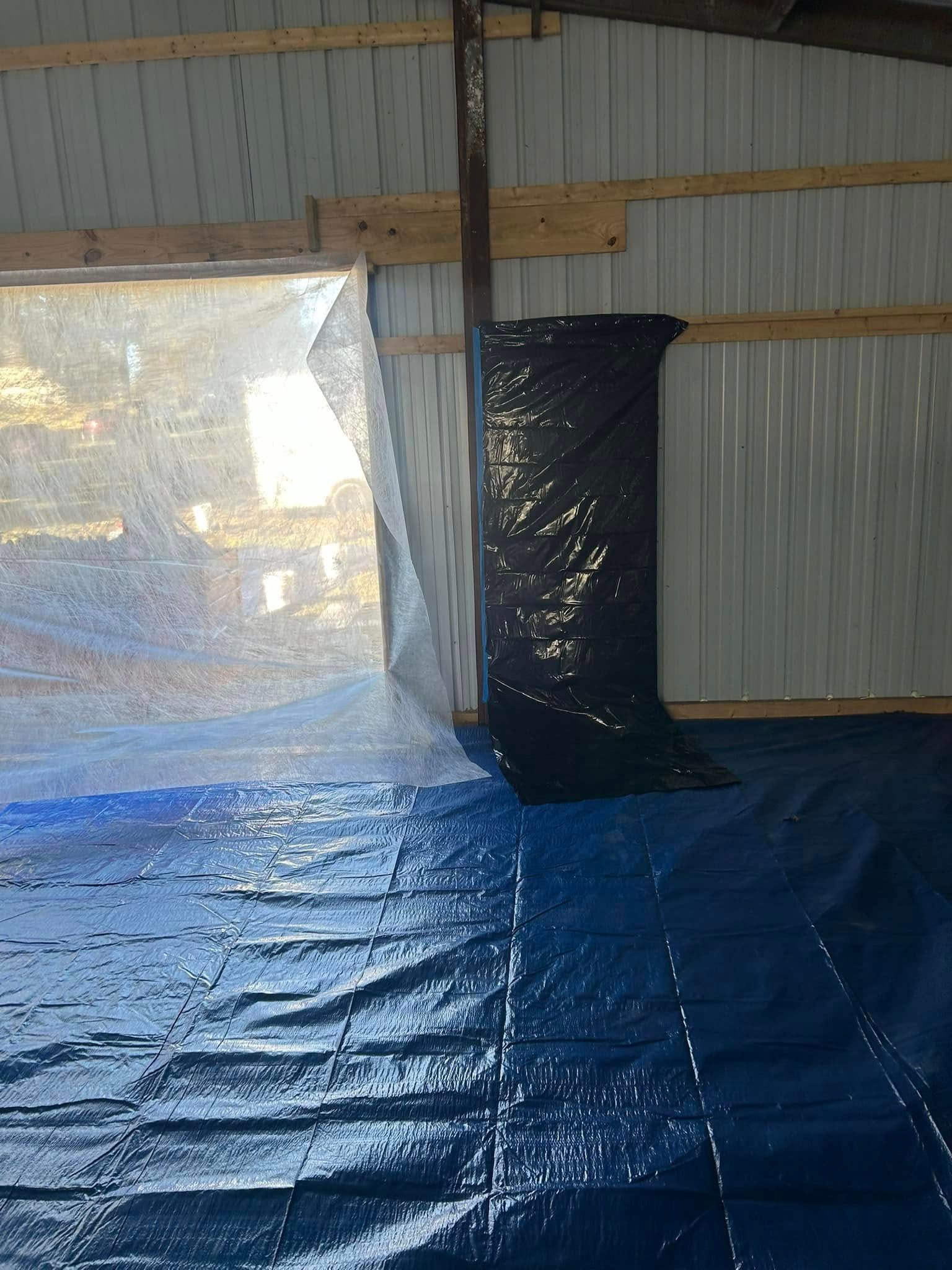 Interior of a building with a blue tarp on the floor and plastic sheeting acting as walls. Black tarp covers a pillar.