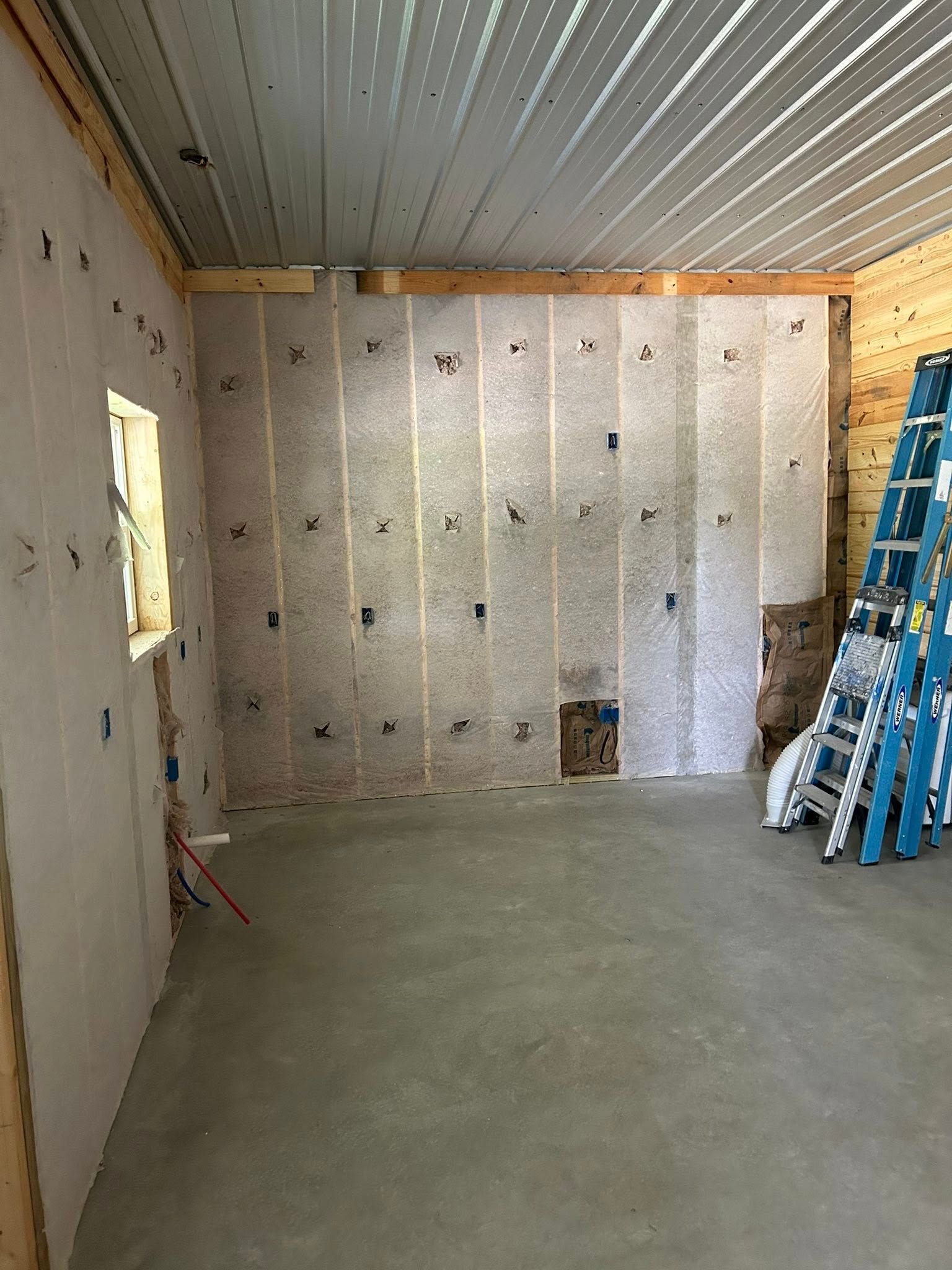 Interior view of a room under construction. Insulation and wiring are visible on the walls and ceiling. A ladder leans against the wall.