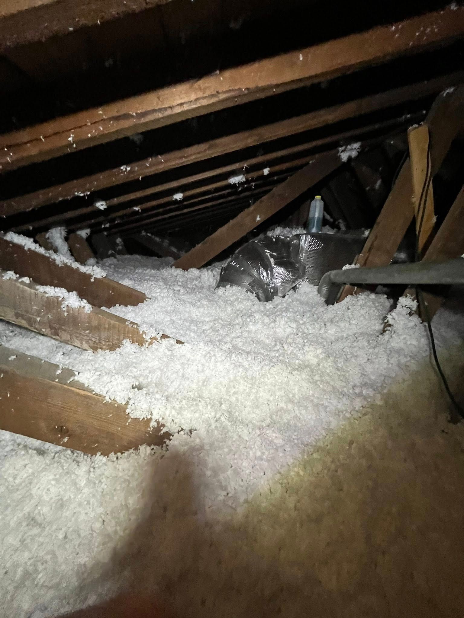 Attic filled with fluffy white insulation between wooden beams. A flexible, silvery duct is visible in the center.