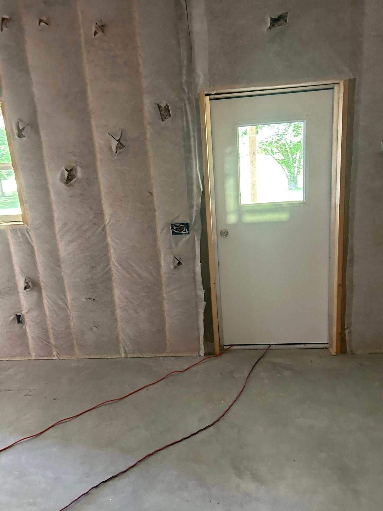 Interior view of a room under construction with insulation on the walls, a white door, and electrical wiring on the concrete floor.