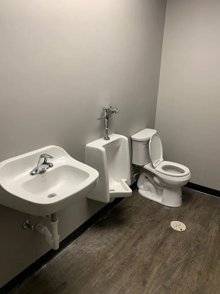 A public restroom with a sink, urinal, and toilet on a gray-walled background. Dark floor.