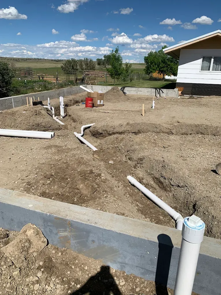 Construction site with white PVC pipes laid in trenches, near a concrete foundation. Bright blue sky.