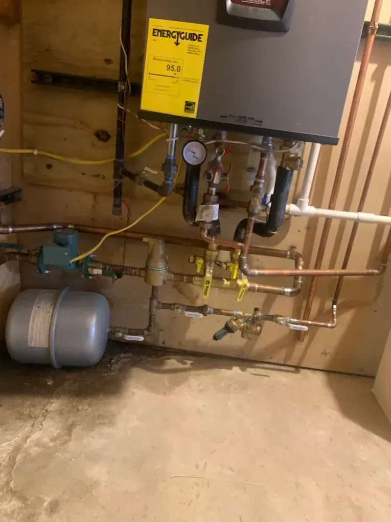 Wall-mounted boiler with copper pipes, valves, and expansion tank in a basement setting.