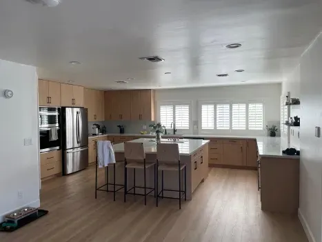 Modern kitchen with light wood cabinetry, a central island with two bar stools, stainless steel appliances, and white walls.