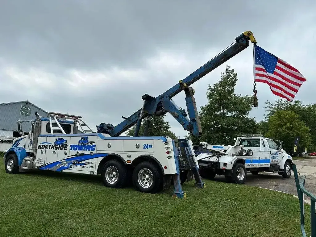 Two tow trucks are parked in a grassy field with an american flag flying in the background.