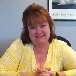 A woman in a yellow shirt is sitting at a table with her hands folded and smiling. A woman in a yellow shirt is sitting at a table with her hands folded and smiling.
