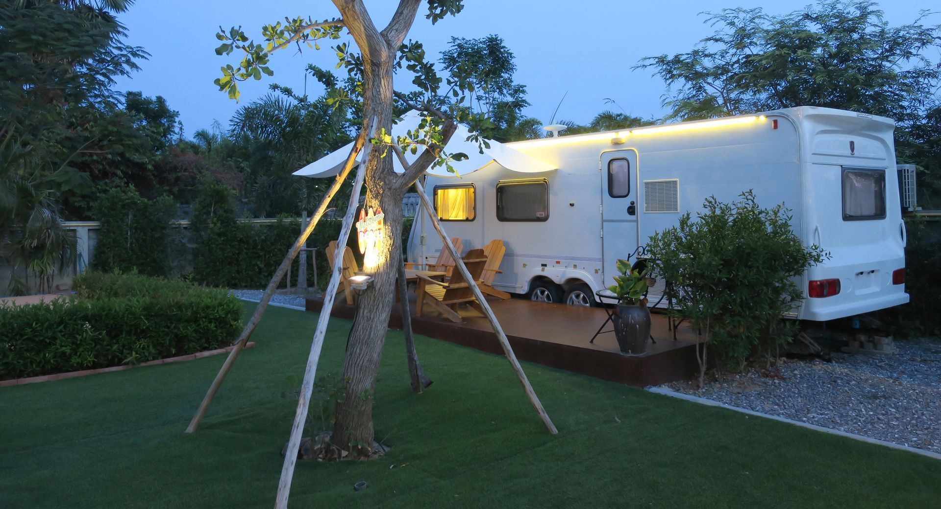 White RV parked on grass, illuminated by lights, next to a small tree. Dusk setting.