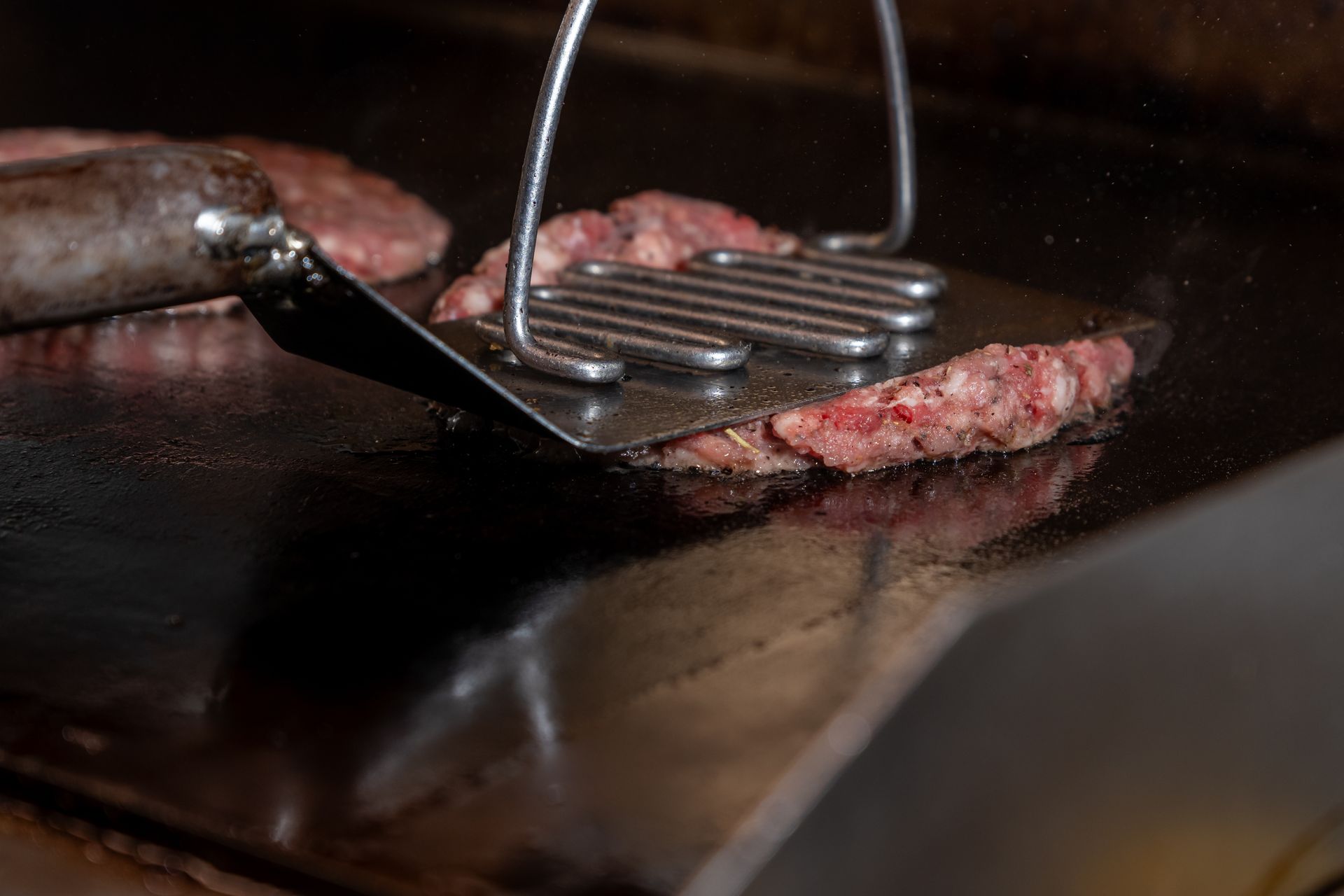 A spatula and a press flatten a raw hamburger patty on a hot, shiny grill.