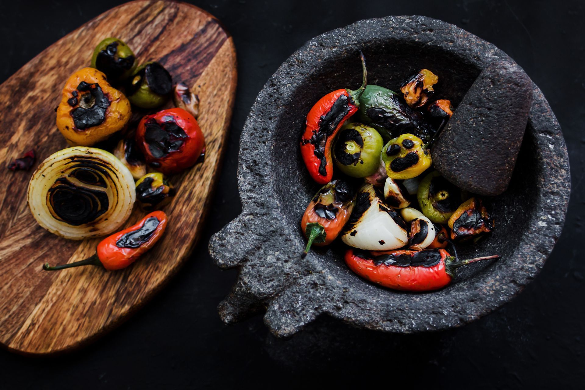Charred vegetables in a mortar and on a wooden board; peppers, onions, and tomatoes.
