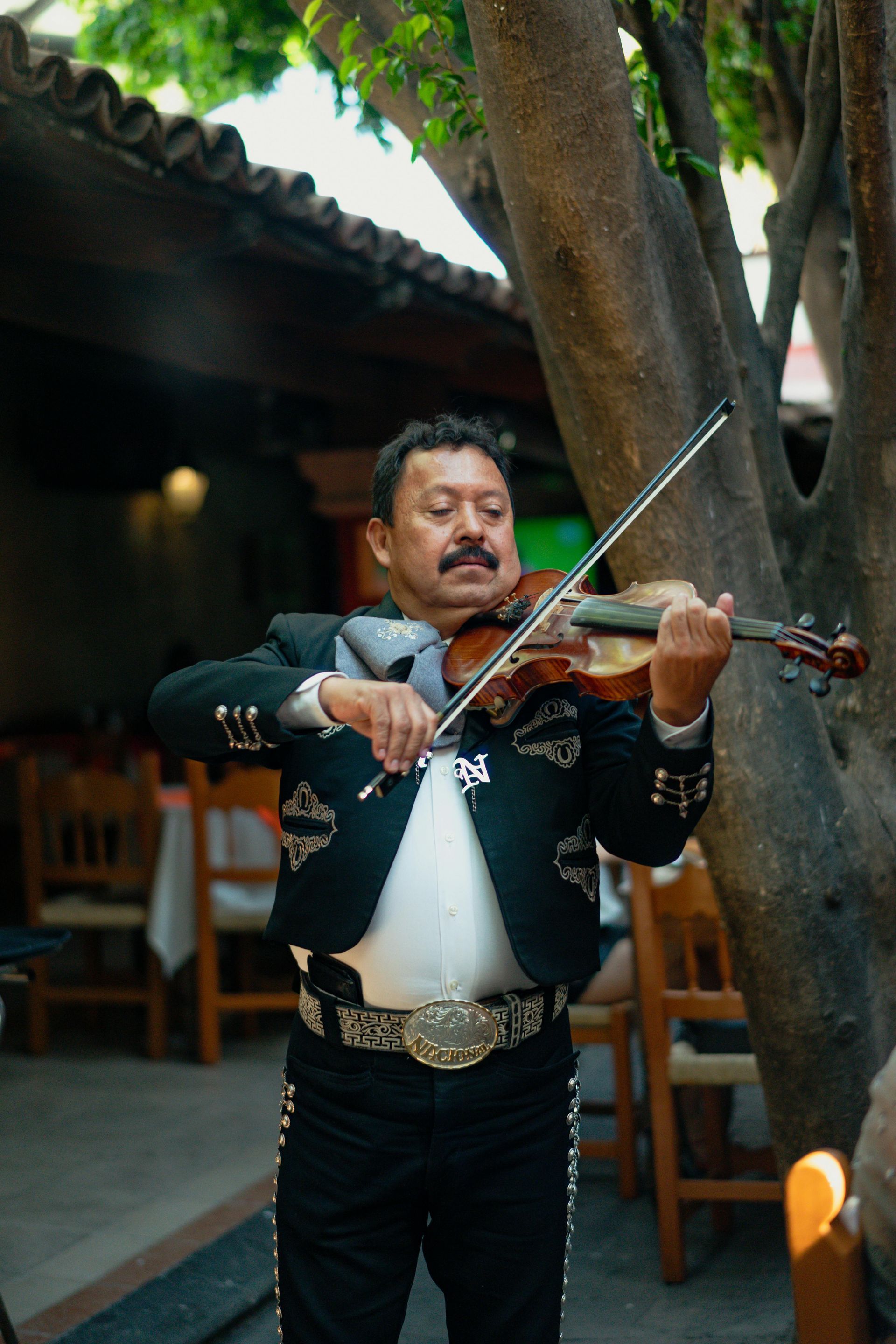 Mariachi musician playing violin outdoors. He wears a black suit, and stands near tables.