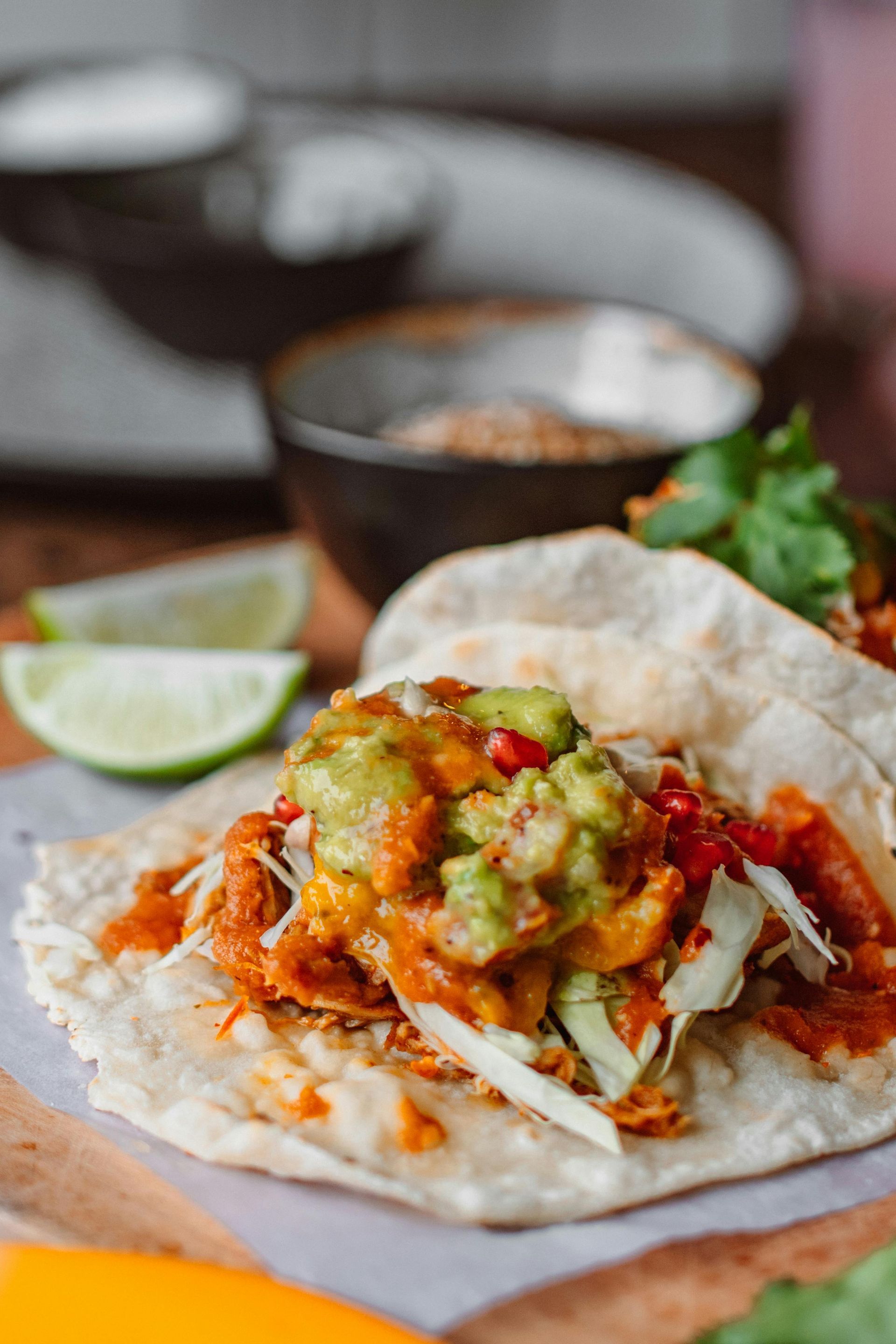 Tacos topped with guacamole, salsa, and shredded cabbage on a wooden board with lime slices and bowls of spices.