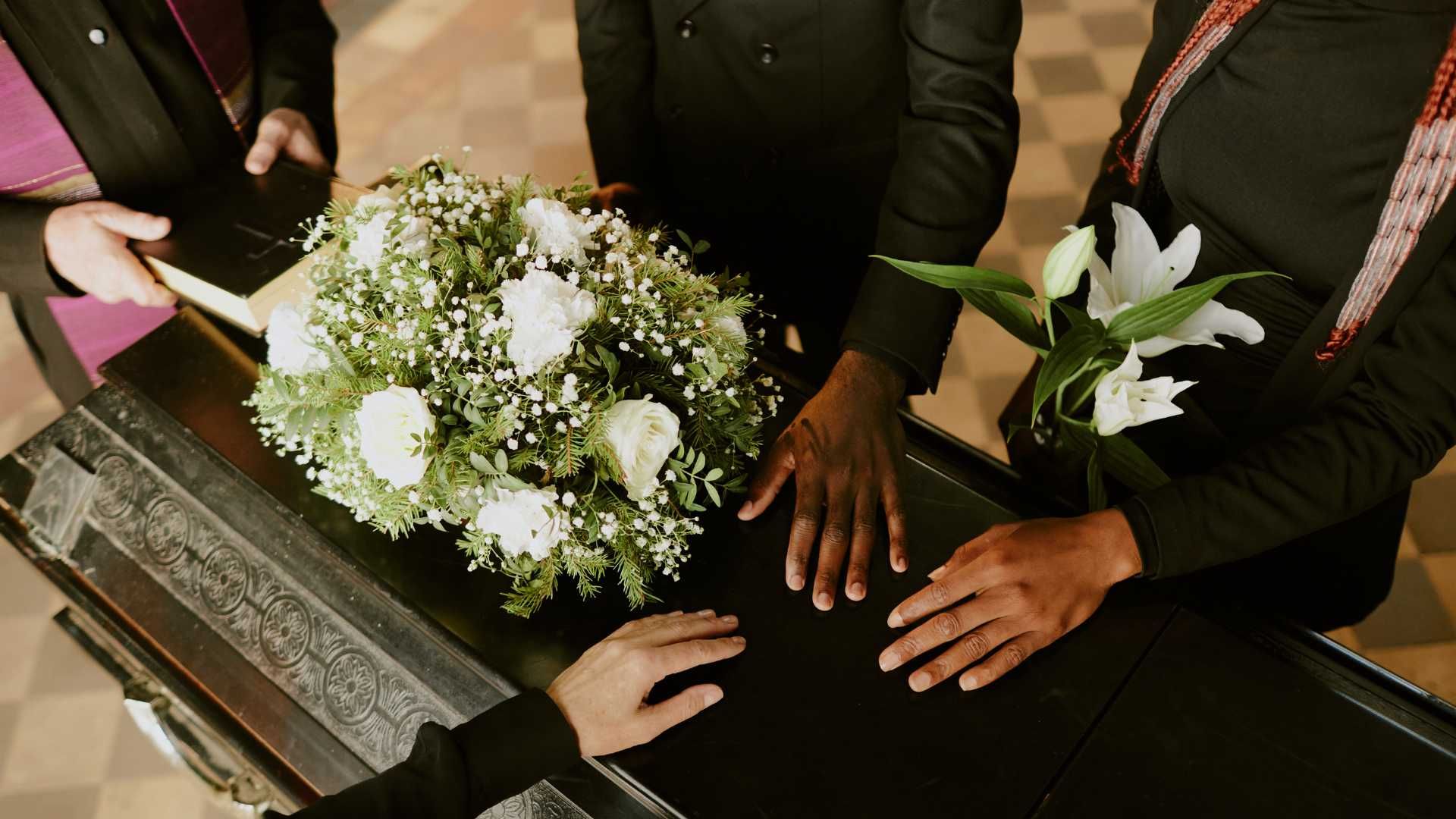 A group of people are standing around a coffin with flowers on it.