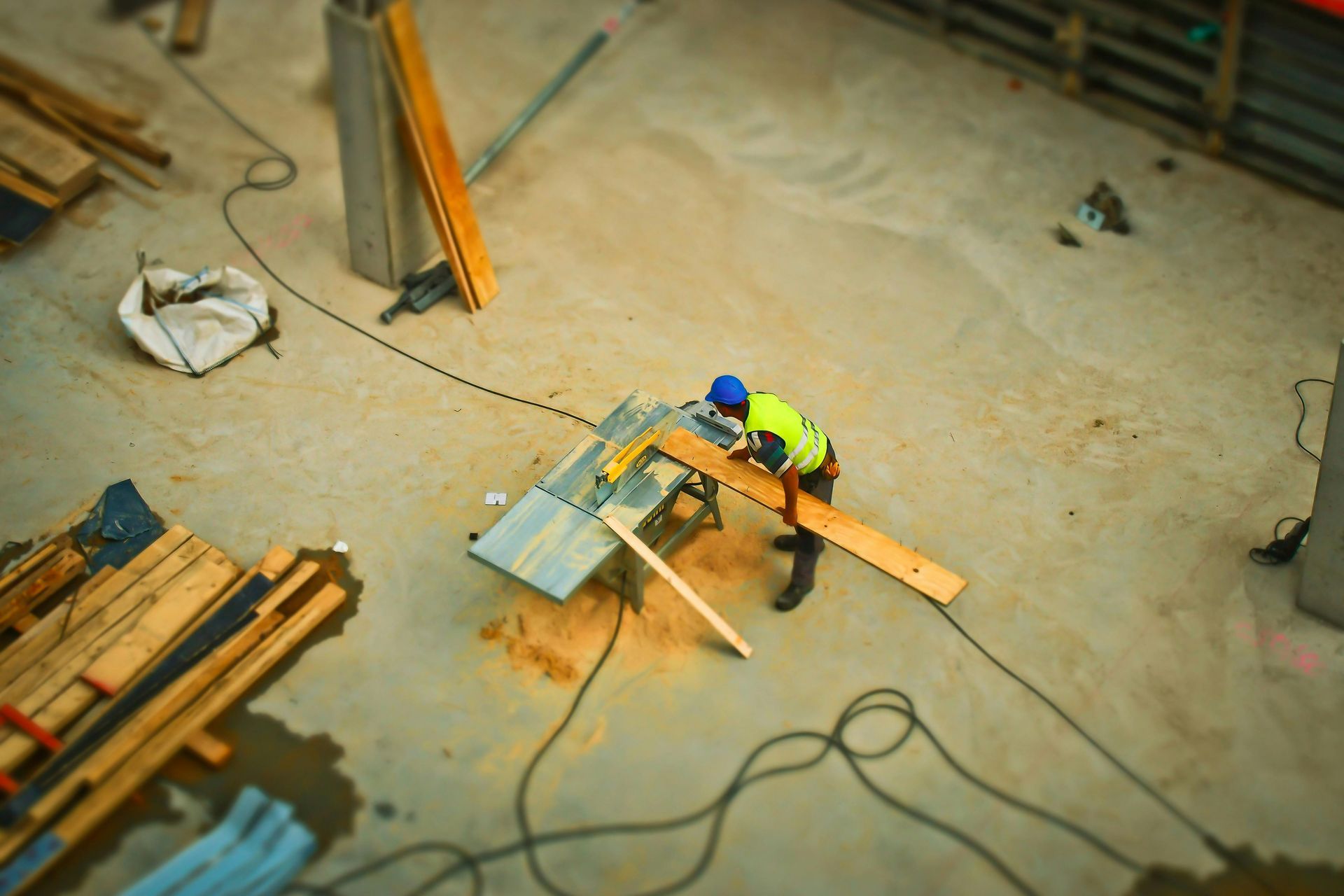 A worker in a yellow safety vest uses a table saw to cut a wooden plank at a construction site.