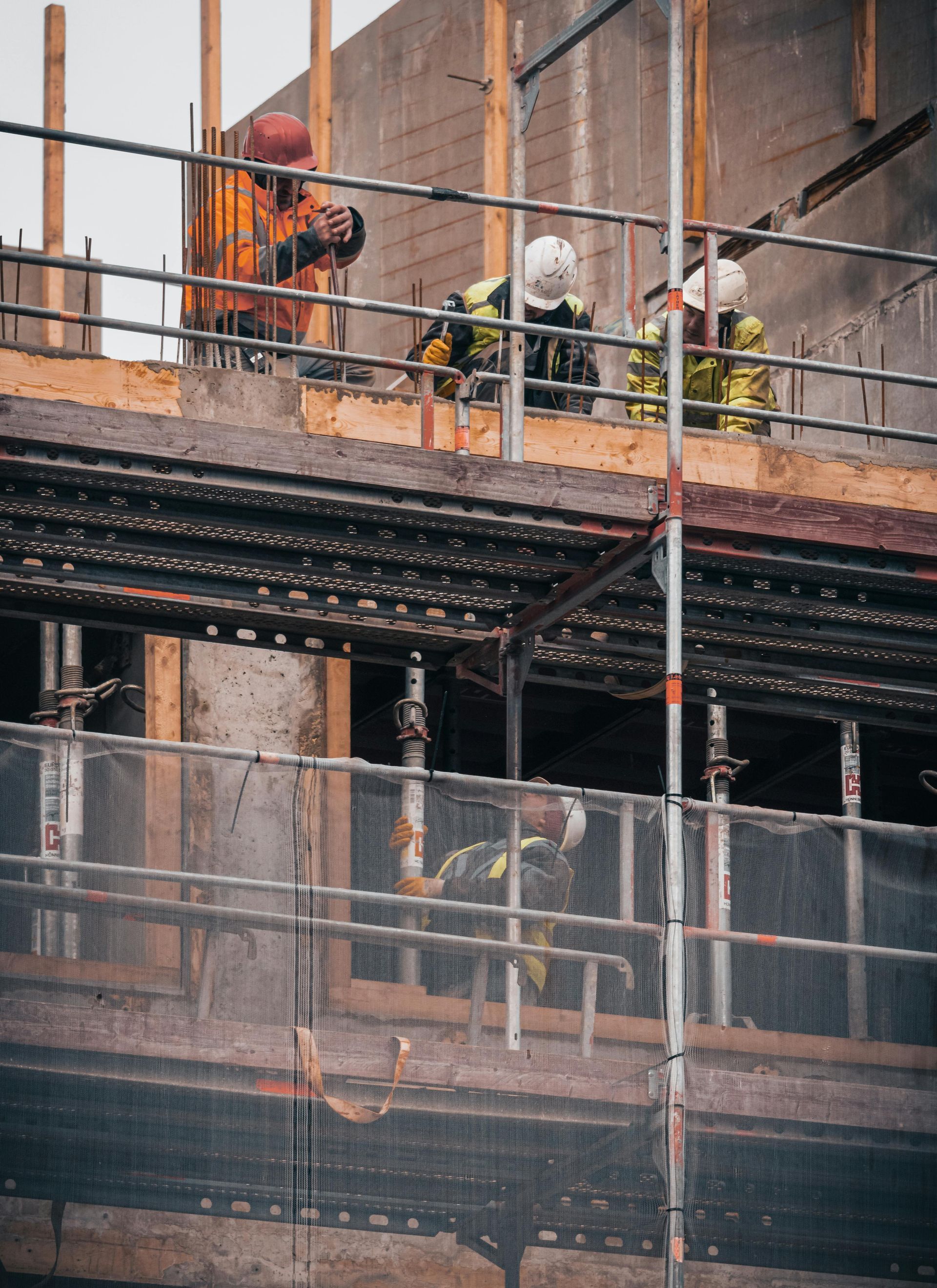 Construction workers in high-visibility gear and hard hats work on a multi-level building site with metal scaffolding.