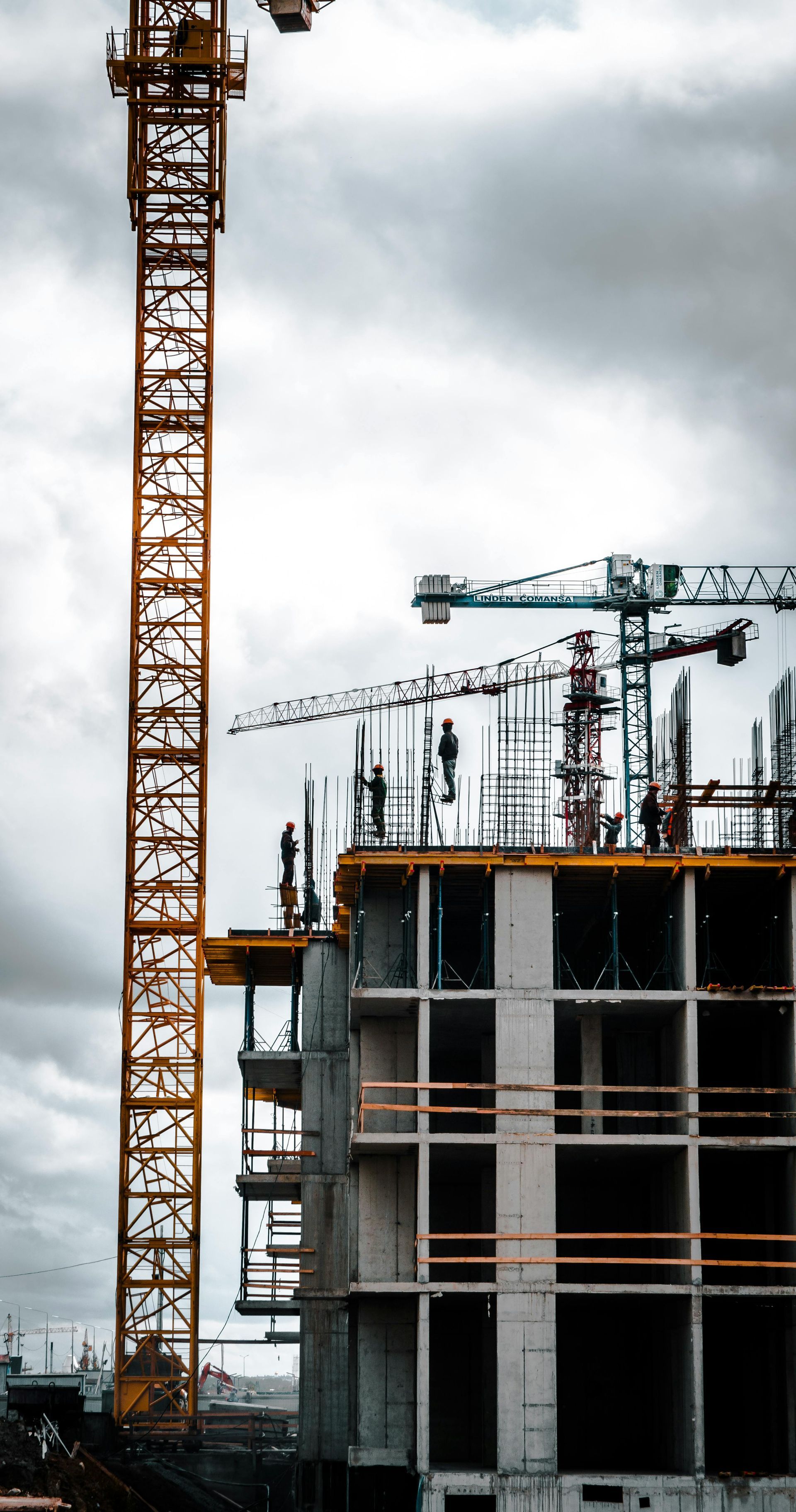 Construction workers on the top level of a building under construction, with two cranes against a cloudy sky.