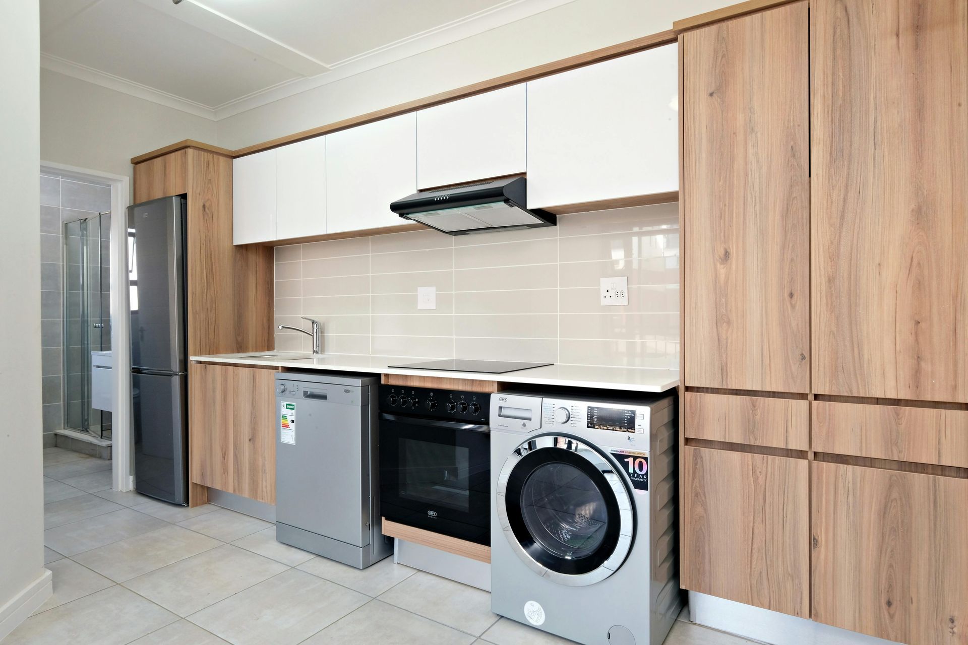 A modern kitchen with light wood cabinets, white upper cabinets, stainless steel appliances, and a tiled backsplash.