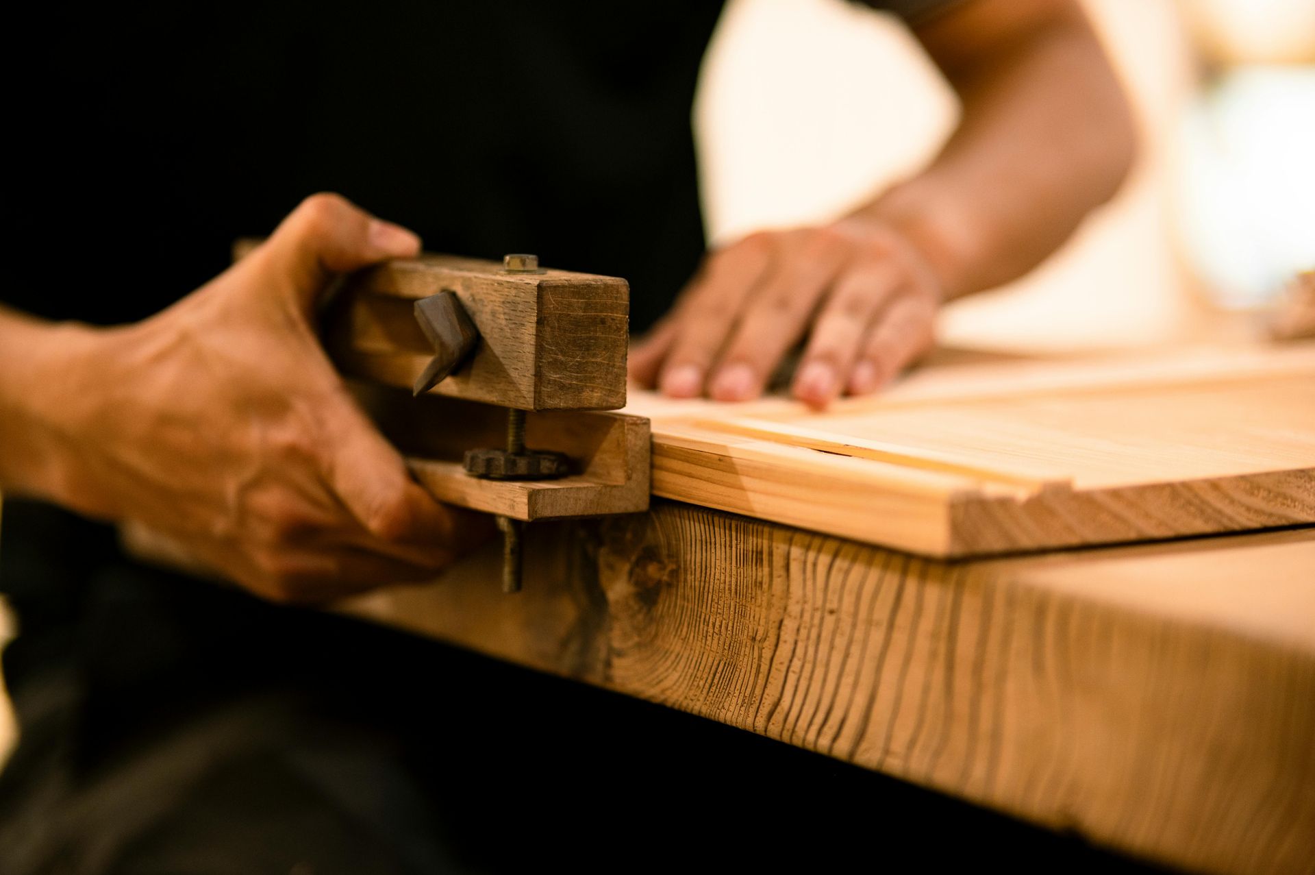 A person uses a wooden marking gauge to measure a wooden board on a workbench.