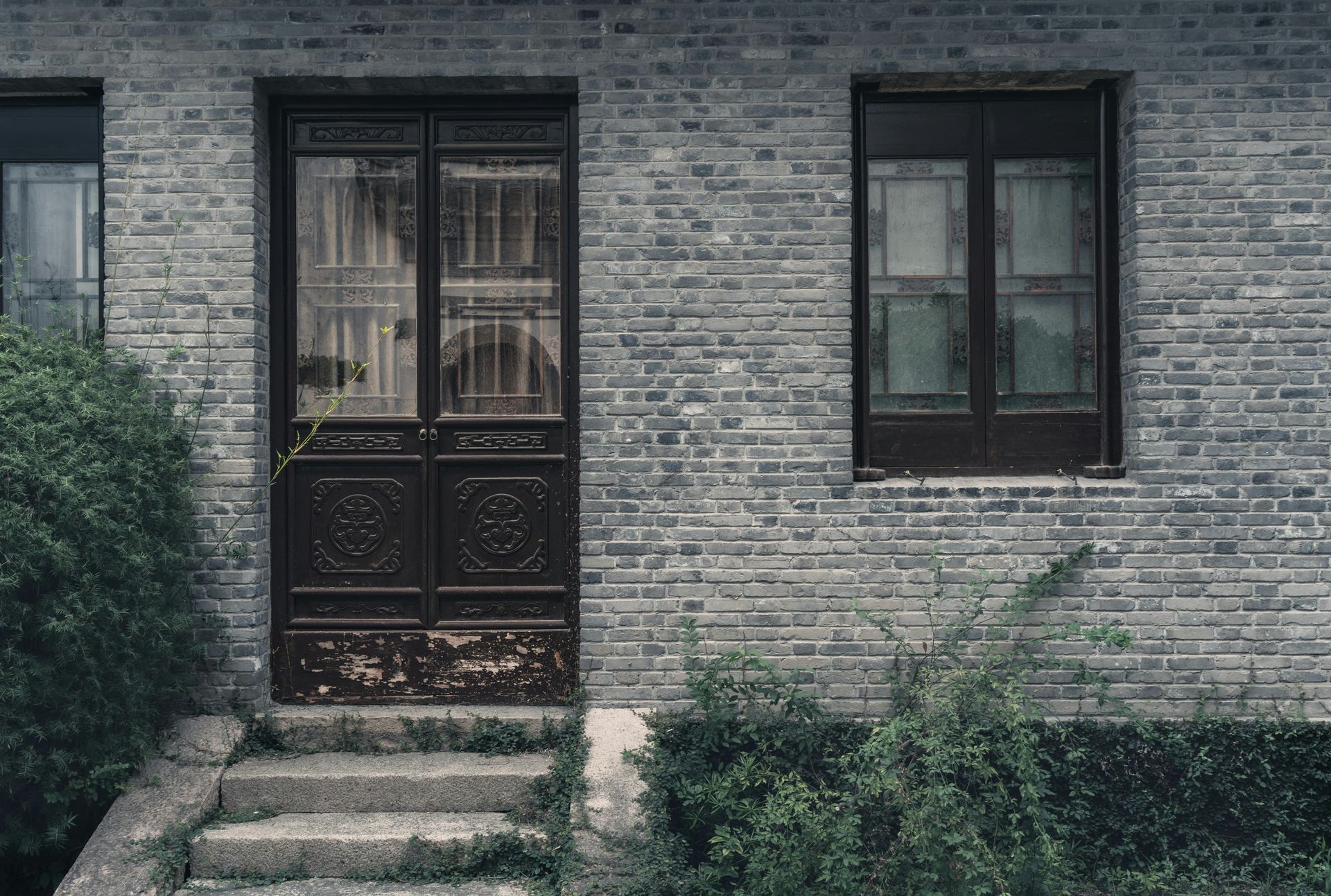 A dark brown double door and a window are centered on a weathered gray brick wall, with stone steps and plants below.