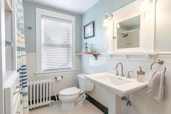 A light blue bathroom featuring a pedestal sink, white tile walls, a toilet, a window with blinds, and a striped shower.
