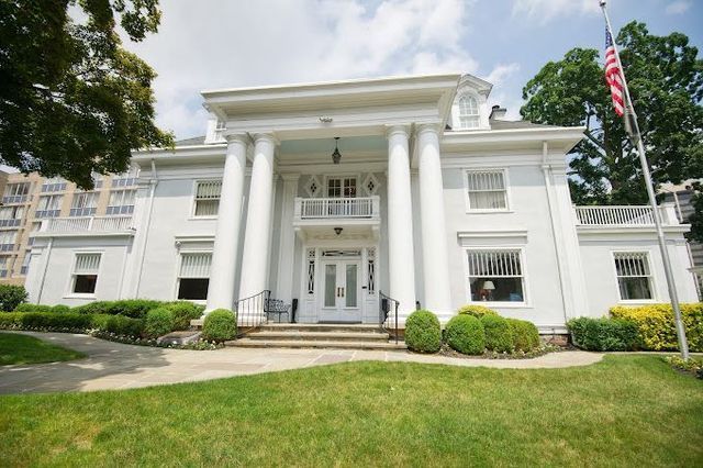 White, two-story funeral home with large columns and an American flag in front. Green lawn and shrubs.