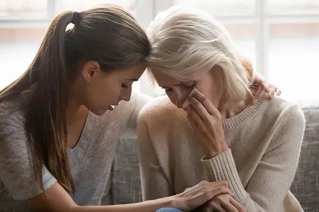 Woman comforts another woman who is crying; both are indoors, close together.
