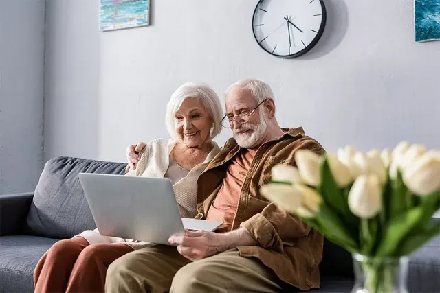 Elderly couple on couch looking at laptop, smiling. Living room with clock and flowers.