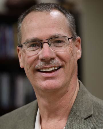 Man wearing glasses, smiling with a mustache, in a blazer.