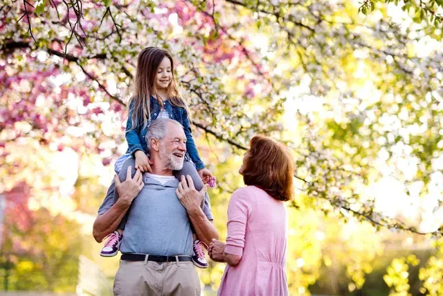 Grandfather carrying a child on his shoulders in a park, smiling. Grandmother holds their hands under pink and yellow flowers.