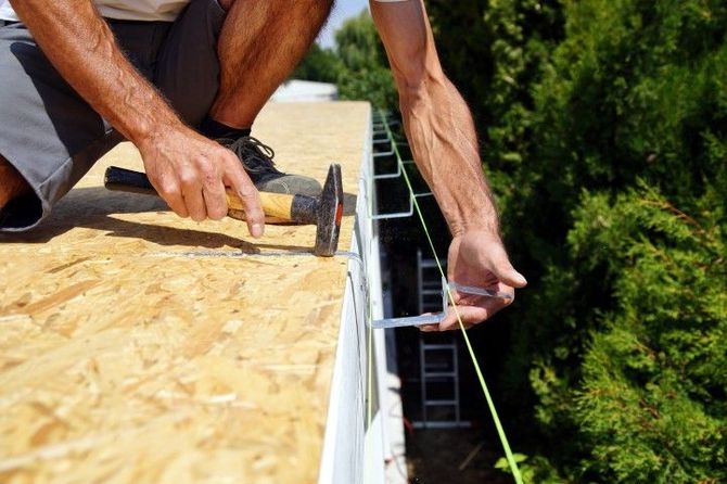 Worker hammering roofing nails on an OSB roof edge with a safety line attached