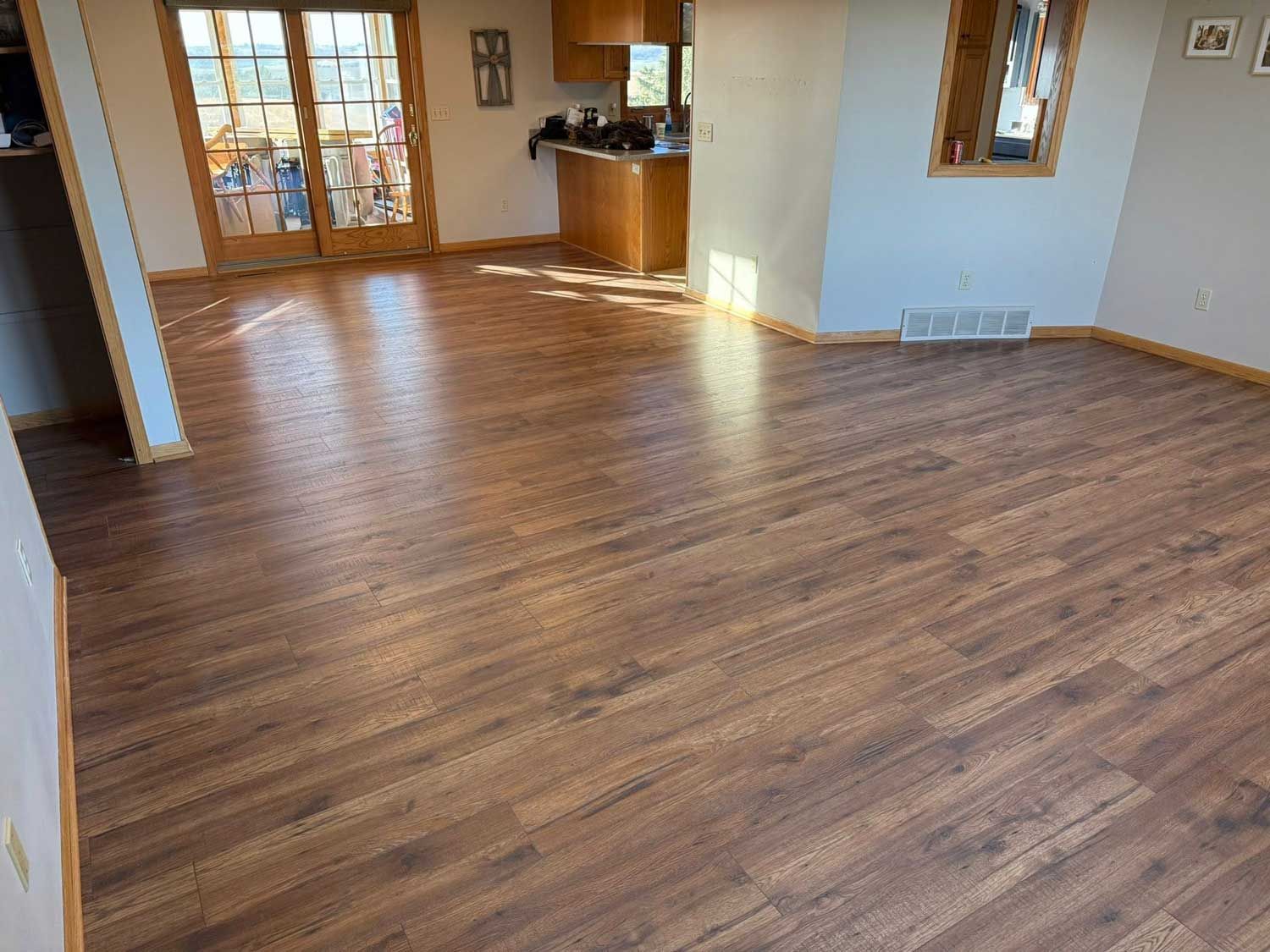 Wooden-floored room with sunlight, french doors, and a small kitchen visible in the background.