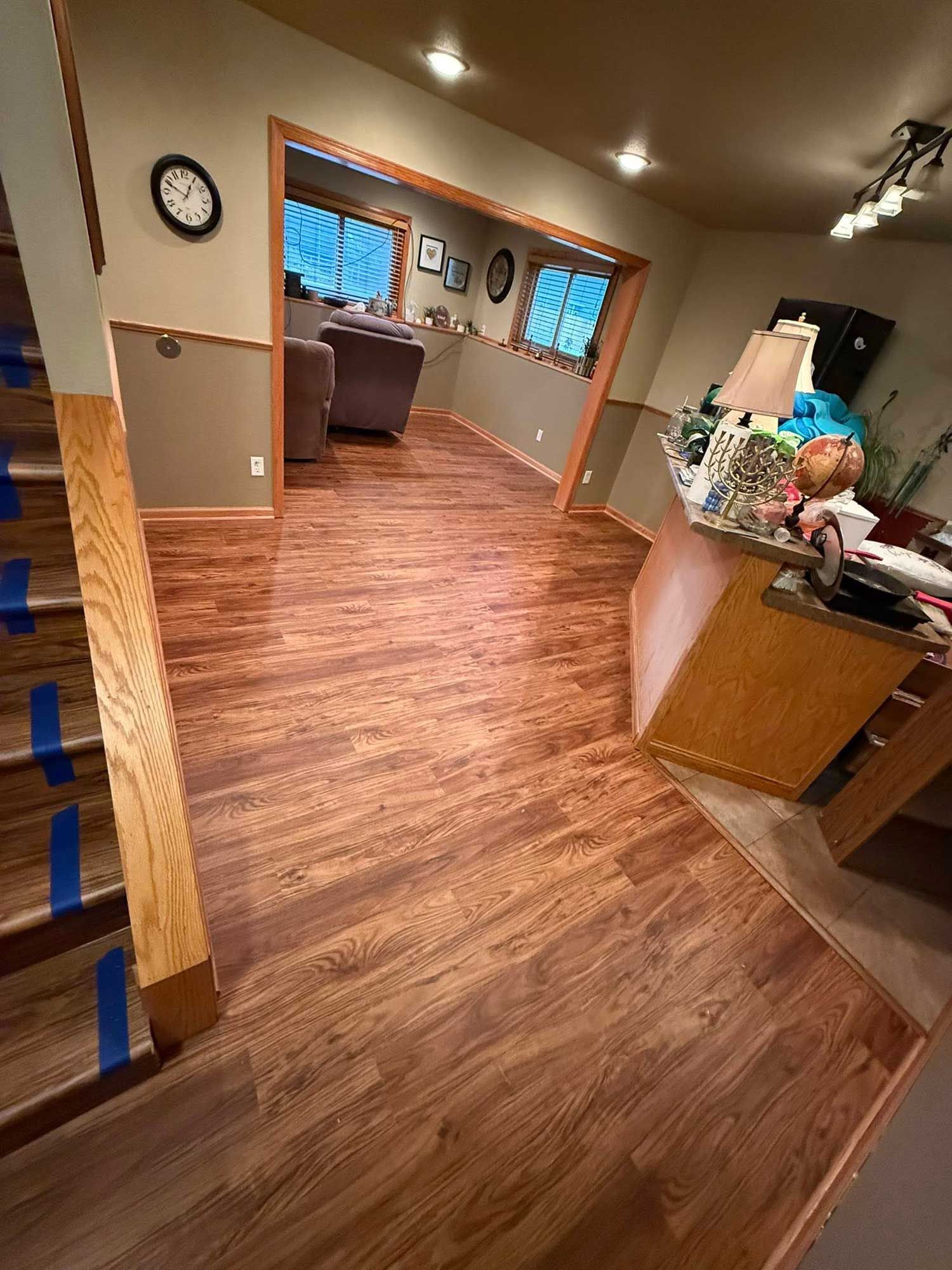 Interior view of a room with wood flooring, stairs, and a partially visible living area with windows.