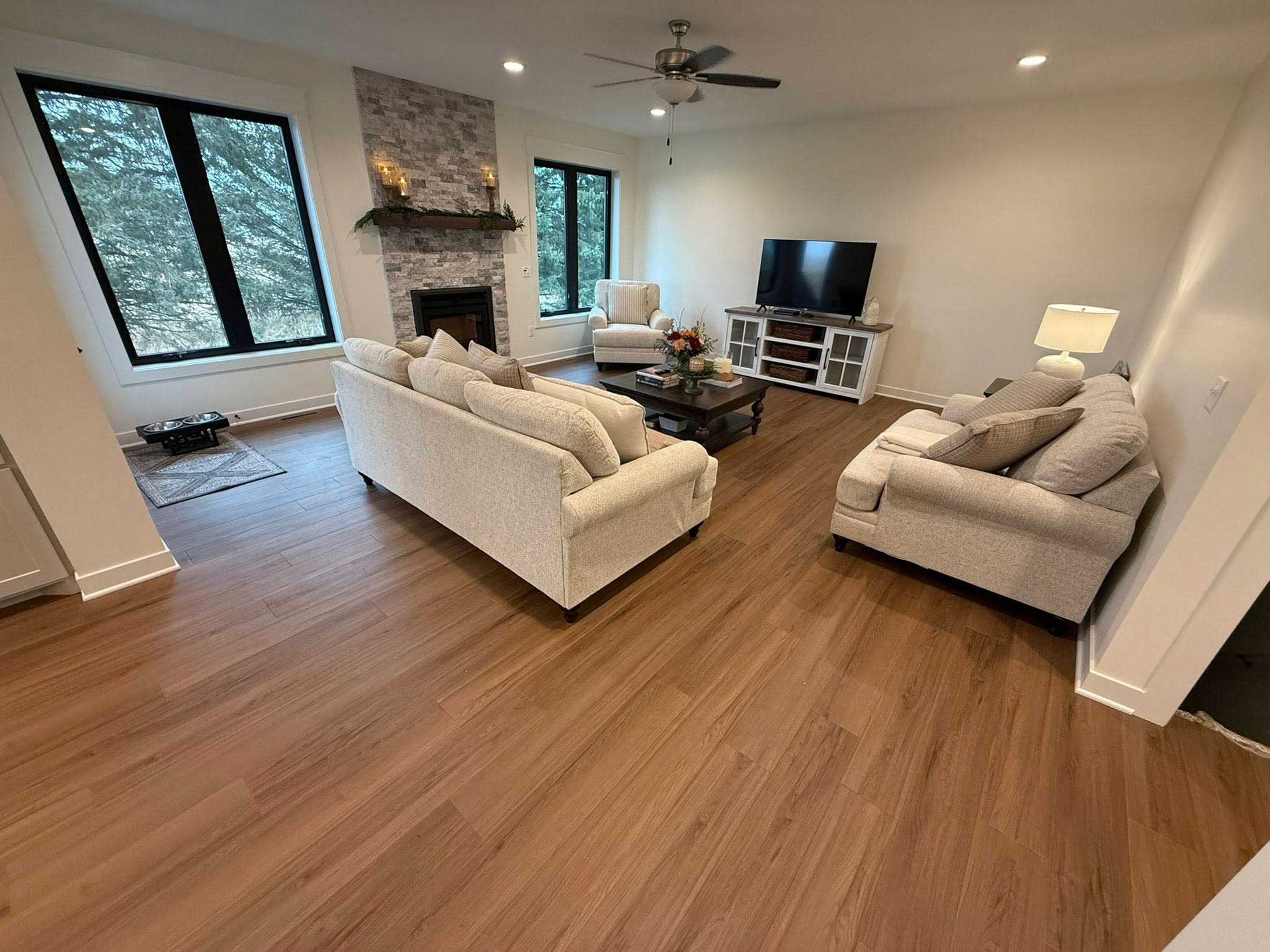 Living room with light-colored sofa and armchair, fireplace, TV, and wood-look flooring. Large windows look out to green trees.
