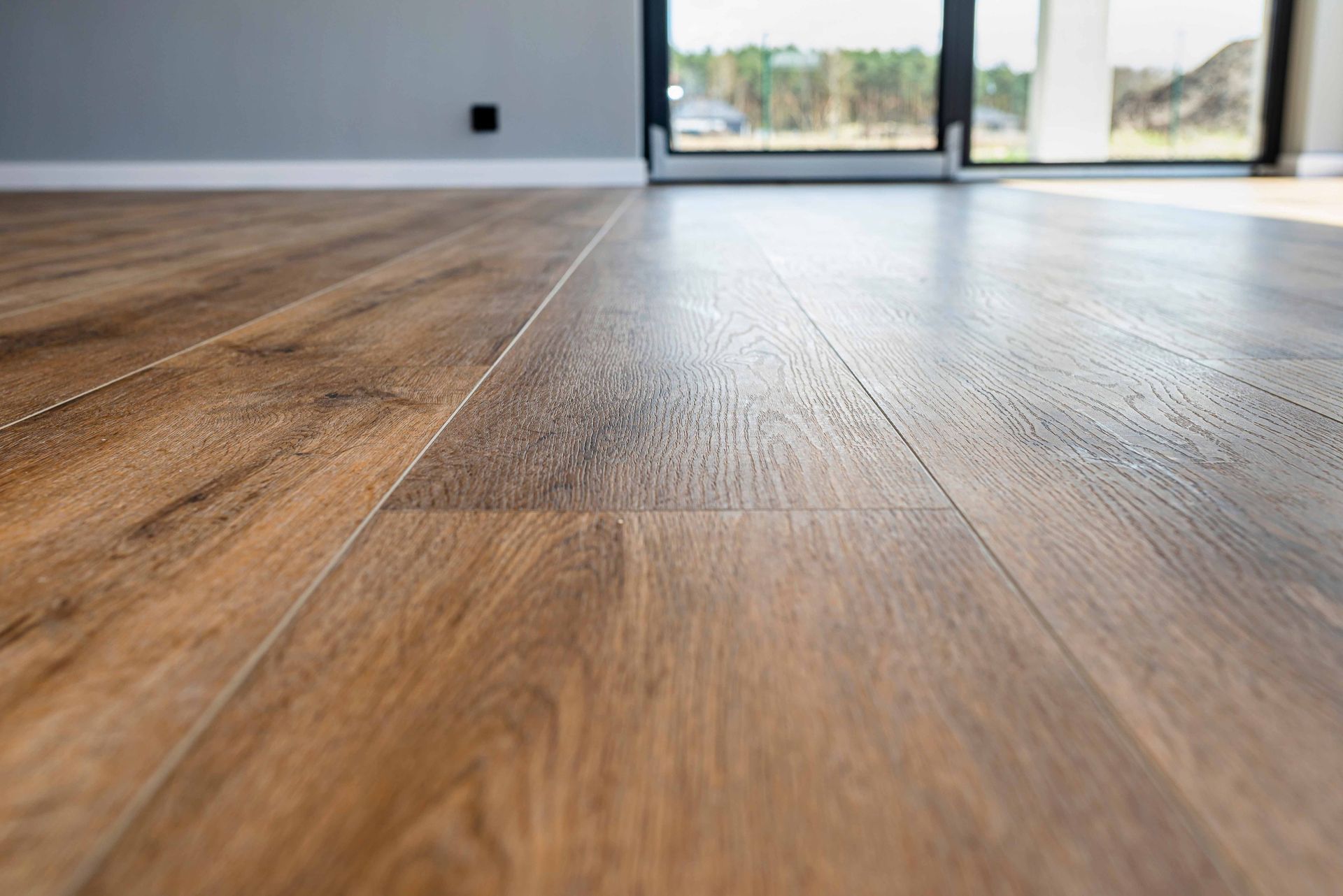 Close-up of glossy wood-look flooring with long planks, leading to a window and gray wall.