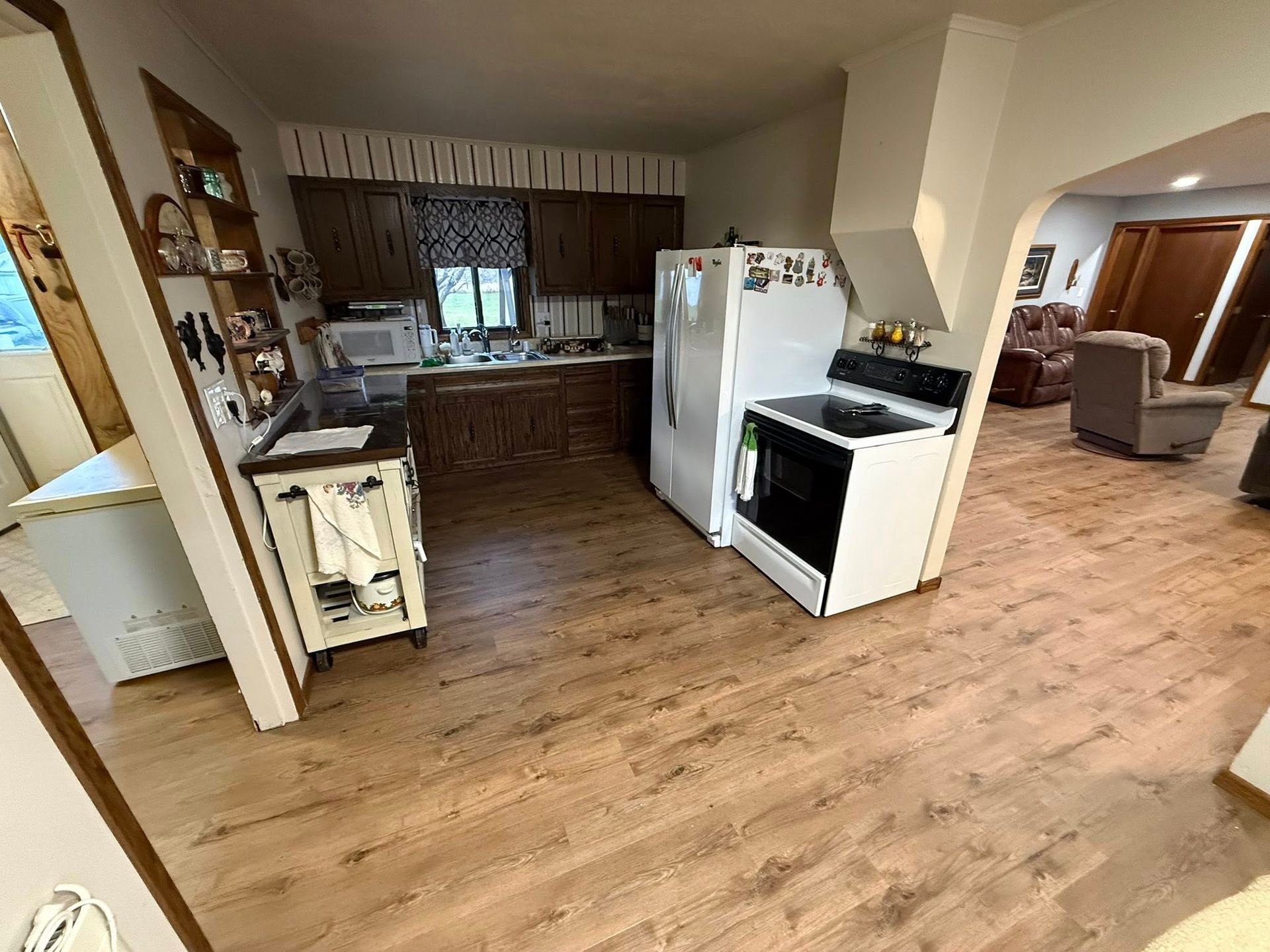Kitchen with brown cabinets, white appliances, and light-colored wood flooring. View into a living area.
