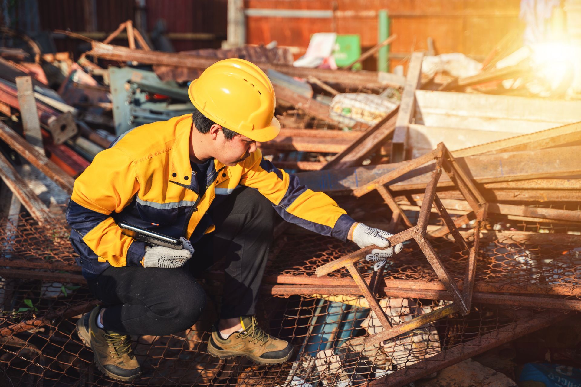 A worker in a yellow hard hat and uniform inspects rusted metal scraps in a salvage yard under bright sunlight.