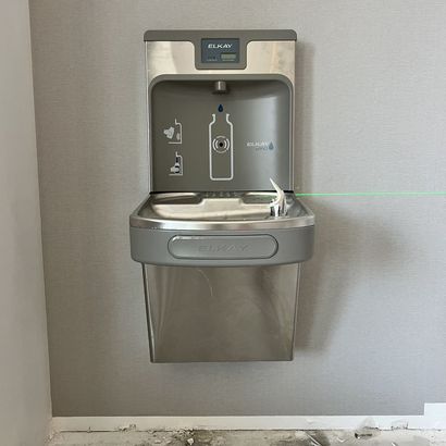 Wall-mounted stainless steel drinking fountain with bottle filler against a gray wall