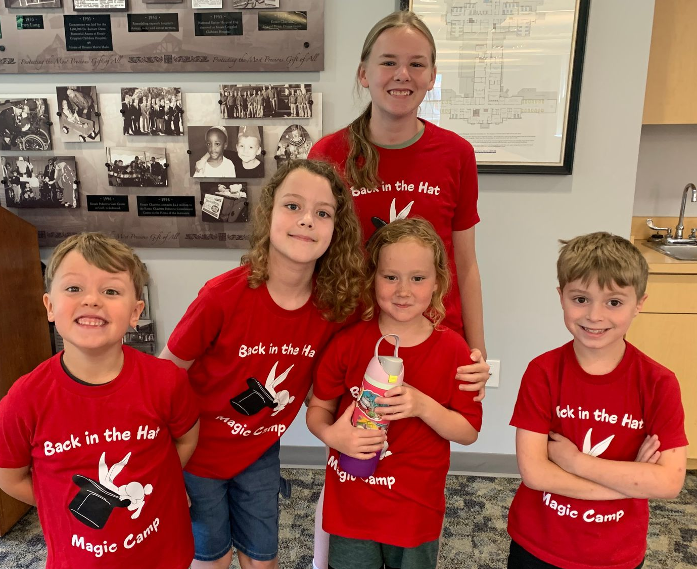 Five children in red shirts posing indoors in front of a bulletin board, one holding a bottle