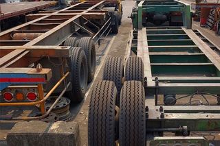 Two semi-trailer chassis in a parking area, one rusty brown, the other green, both with multiple tires.