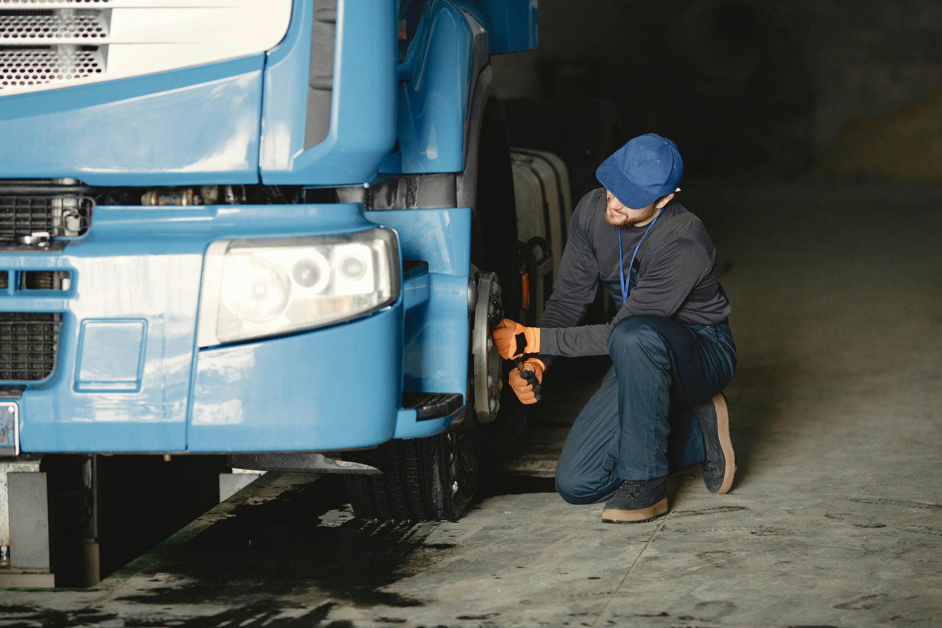 Mechanic inspecting the front wheel of a blue truck in a dimly lit garage. He kneels, wearing a cap and gloves.