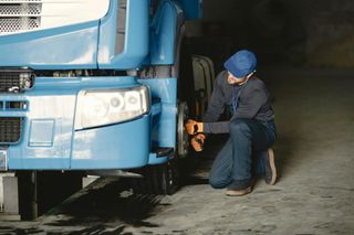 Mechanic inspecting the front wheel of a blue truck in a dimly lit garage. He kneels, wearing a cap and gloves.